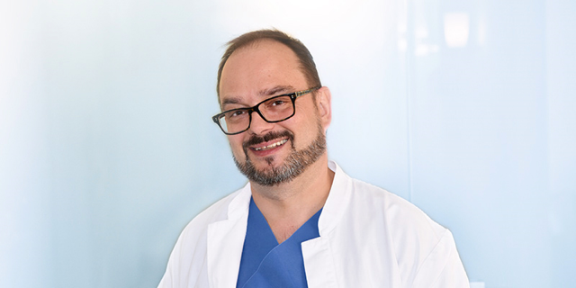 Dr. med. Roger Gmür, lächelnd im weißen Kittel vor hellblauem Hintergrund
