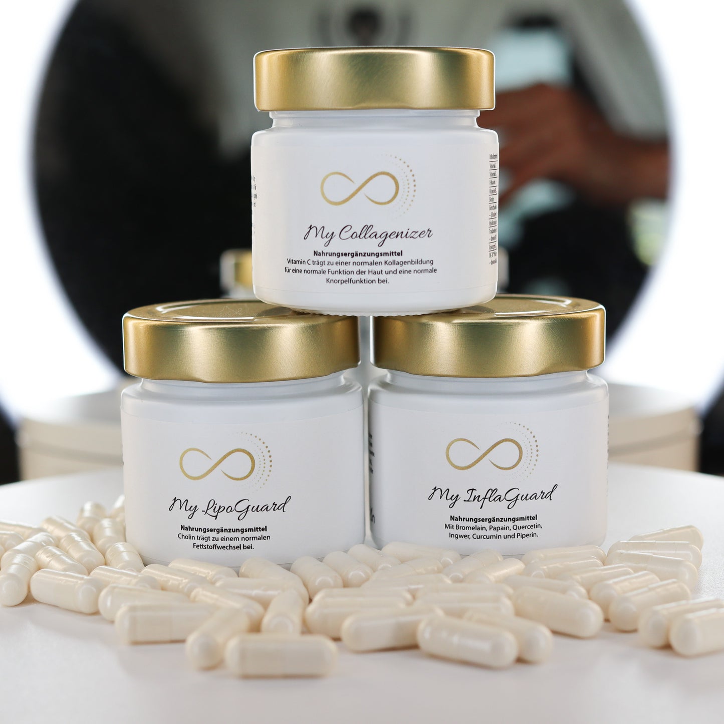 Drei elegante weiße Glastiegel mit goldfarbenem Deckel, beschriftet mit den Produktnamen 'My Collagenizer', 'My LipoGuard' & "My InflaGuard" , stehen dekorativ vor einem Spiegelringlicht. Vor den Tiegeln liegen weiße Kapseln. Hochwertiges Nahrungsergänzungsmittel-Design mit edlem, minimalistischen Look.