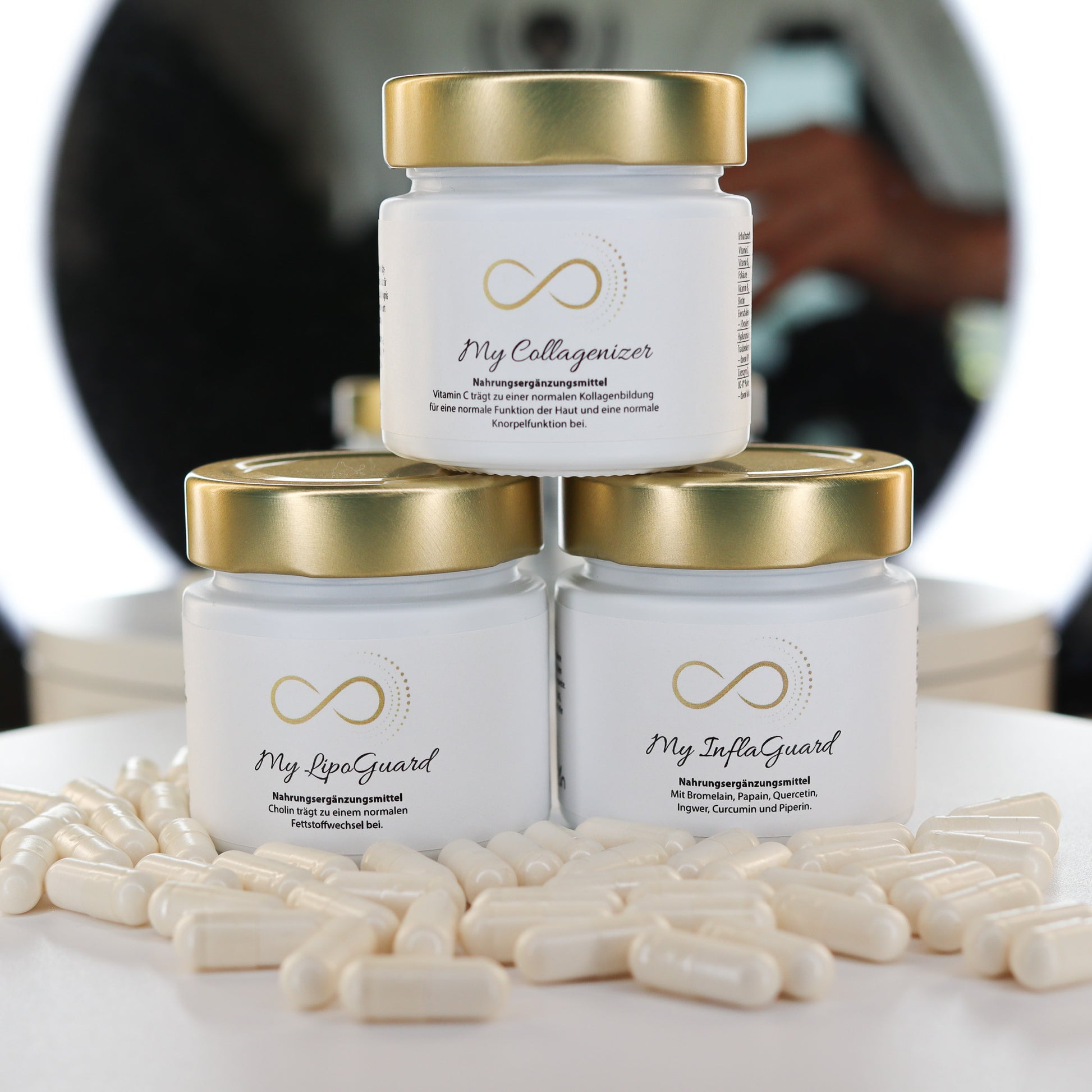 Drei elegante weiße Glastiegel mit goldfarbenem Deckel, beschriftet mit den Produktnamen 'My Collagenizer',  'My LipoGuard' & "My InflaGuard" , stehen dekorativ vor einem Spiegelringlicht. Vor den Tiegeln liegen weiße Kapseln. Hochwertiges Nahrungsergänzungsmittel-Design mit edlem, minimalistischen Look.
