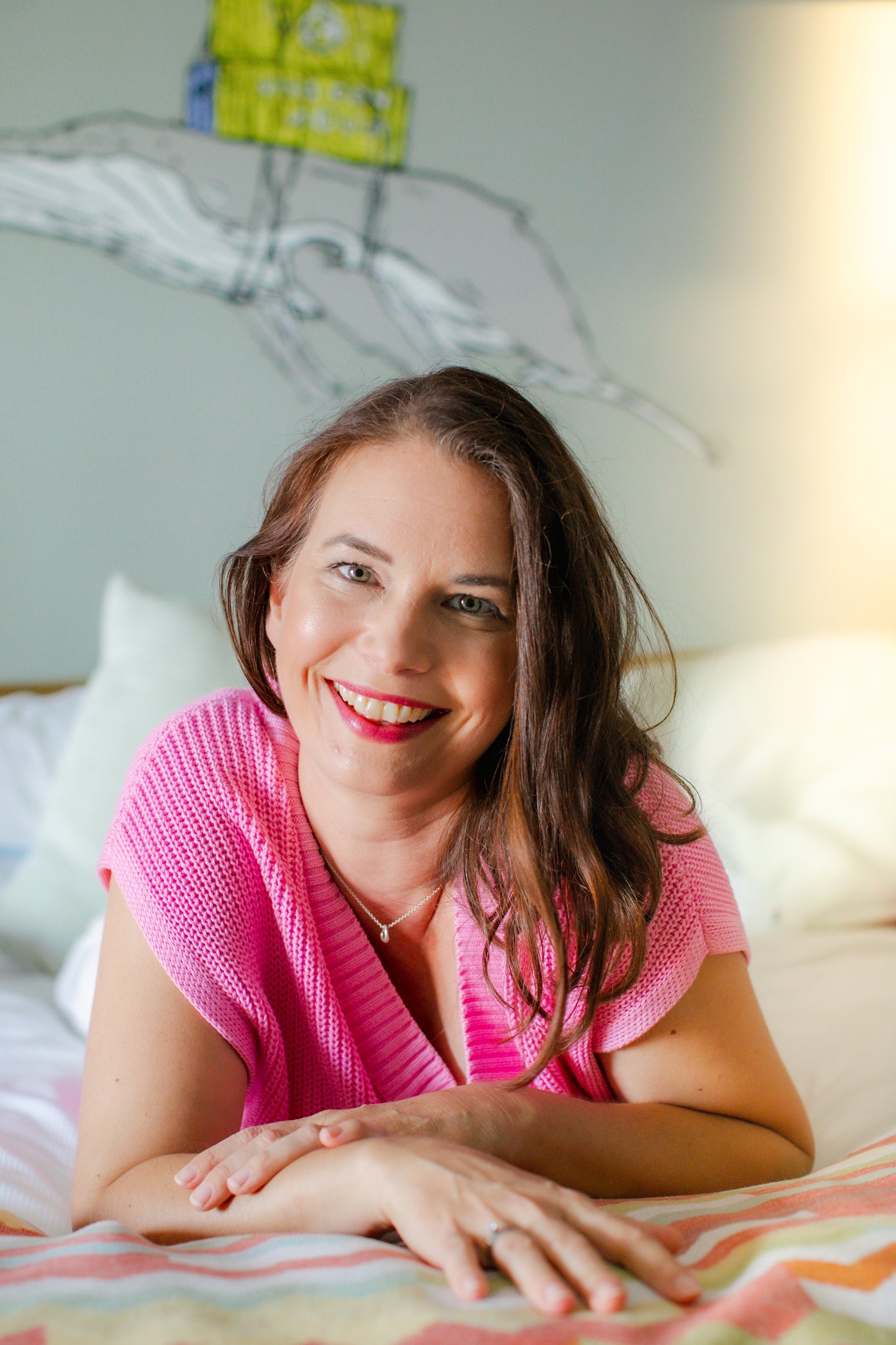 Fröhliche Frau mit langen braunen Haaren und pinkem Strickpullover liegt entspannt auf einem Bett und lächelt in die Kamera. Im Hintergrund stilisierte Zeichnung einer Hand mit einem Haus. Die Atmosphäre wirkt freundlich, offen und einladend.