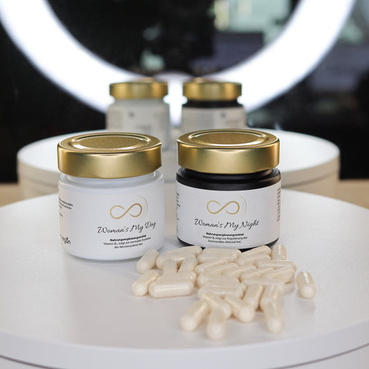 Produktfoto zweier Nahrungsergänzungsmittel aus dem ANS Nutrivity Secrets Sortiment: Woman’s My Day in einem weißen Glastiegel und Woman’s My Night in einem schwarzen Glastiegel, beide mit goldfarbenem Deckel. Die Produkte sind auf einem hellen Präsentationstisch dekorativ neben weißen Kapseln platziert. Die Auswahl basiert auf einer Empfehlung von Katja Rückemann. Detaillierte Angaben zu Inhaltsstoffen und Anwendung befinden sich auf den jeweiligen Produktseiten im Shop.